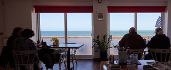 CAFE 1 This image shows the interior of Cafe 1, located in Devon, England, United Kingdom, with people seated at tables near large windows that offer a clear view of the sea. The photograph is taken in the afternoon during early June, indicating a late spring season. The type of photograph is an indoor scene that combines elements of still life and social documentary. The scene captures people enjoying drinks and food inside the cafe, with the blue sea visible outside, suggesting a scenic coastal location. The decor includes plants and a vase of flowers on the tables, while natural light streams in through the windows, highlighting the relaxed atmosphere and the presence of people inside the cafe.